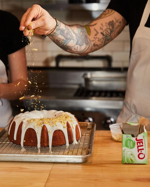Ring cake on a metal cooling rack being decorated by a tattooed arm with sprinkles with Jell-O pudding mix on a wood table.