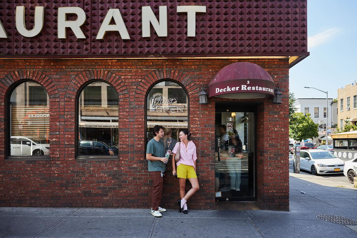 Two people stand outside Decker Restaurant, a brick building with arched windows, under a 'Restaurant' sign on a sunny day in an urban setting.