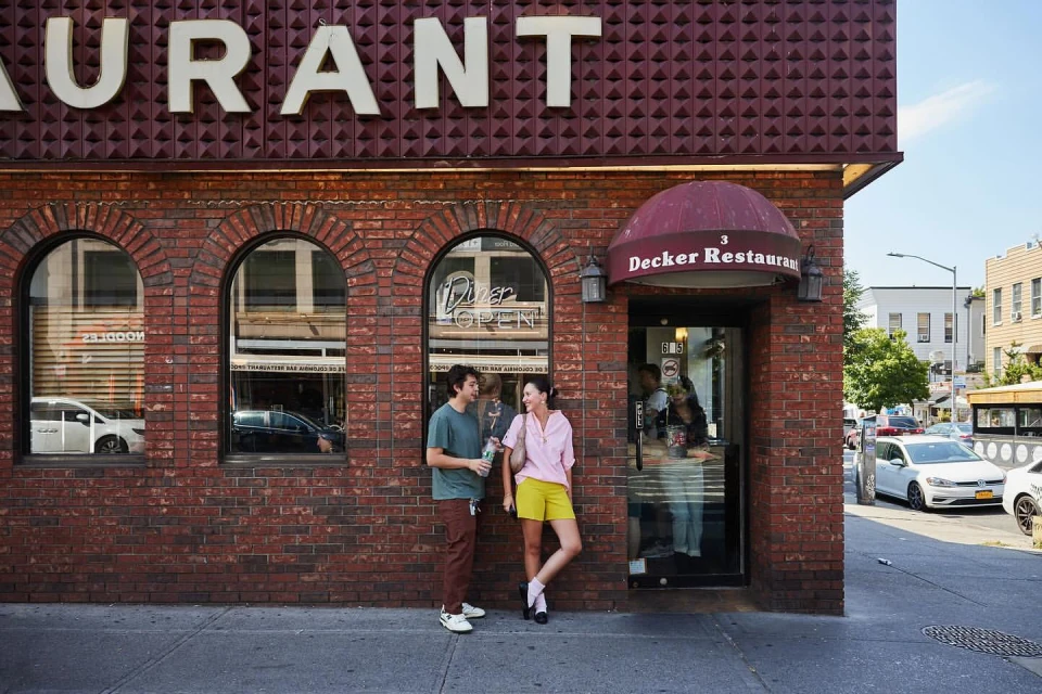 Two people stand outside Decker Restaurant, a brick building with arched windows, under a 'Restaurant' sign on a sunny day in an urban setting.