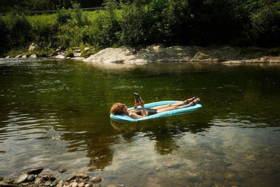 Woman in swimsuit floating on a blue raft in a river, using a cell phone, surrounded by green trees, rocks, and natural light.