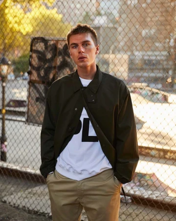 Portrait of a man in an olive jacket, white graphic t-shirt, and khaki trousers standing outdoors near a chain-link fence.
