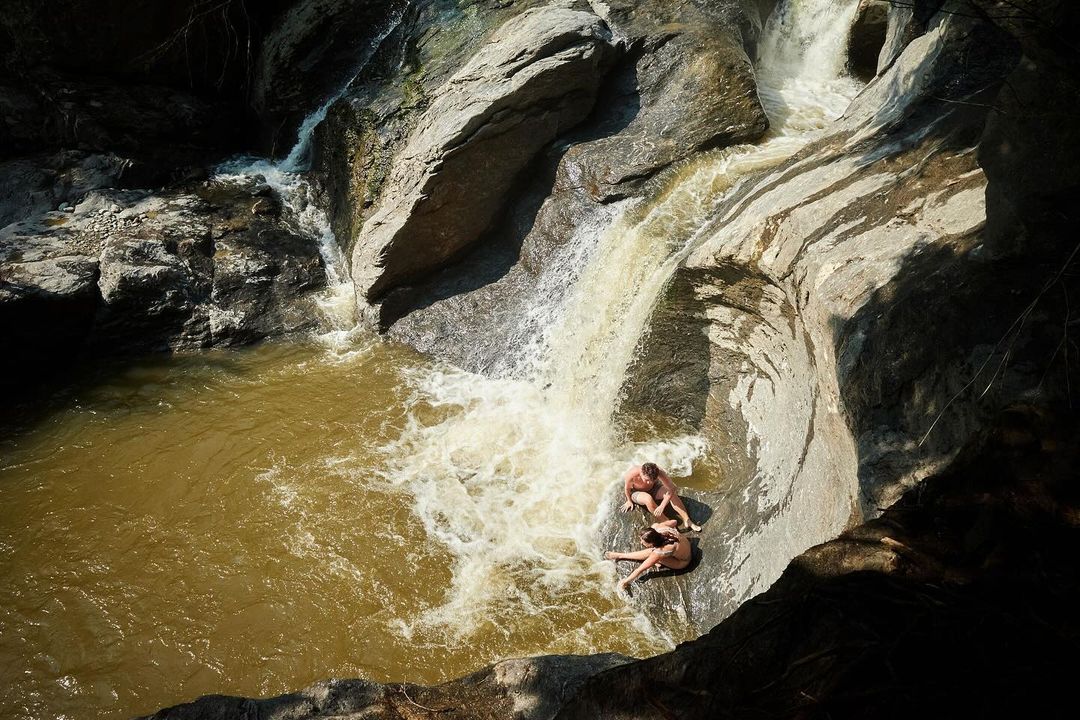 Two adults are sitting at the base of a natural rock waterslide formed by a waterfall flowing into a pool of water in a rocky environment.