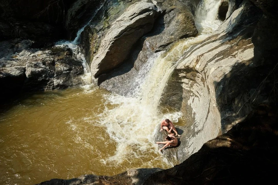 Two adults are sitting at the base of a natural rock waterslide formed by a waterfall flowing into a pool of water in a rocky environment.