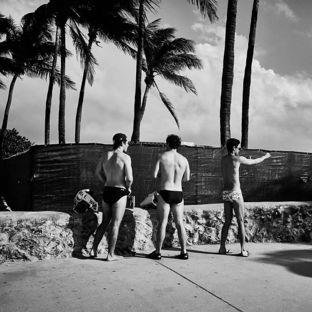 Three men in swimsuits stand near a stone wall with palm trees and a cloudy sky visible in the background in a black and white outdoor shot.