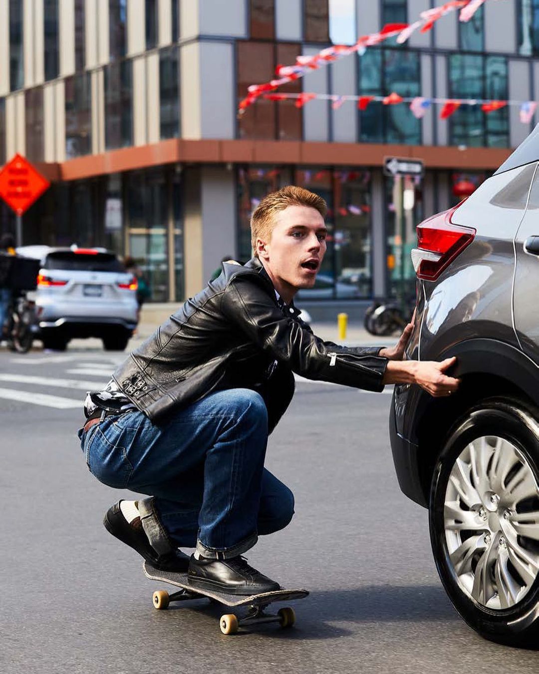 Adult male riding a skateboard on a city street, with a car in the foreground and buildings and banners in the background during the day.