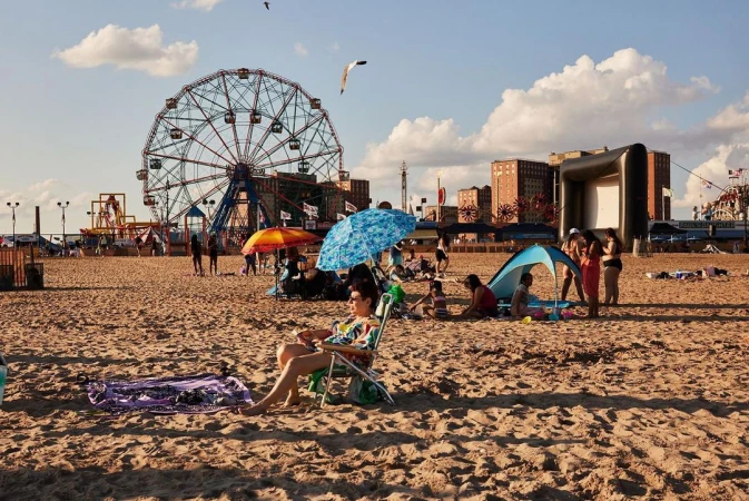 Beach scene features woman relaxing in chair with Ferris wheel, clouds, beachgoers, tents, and various summer activities visible.