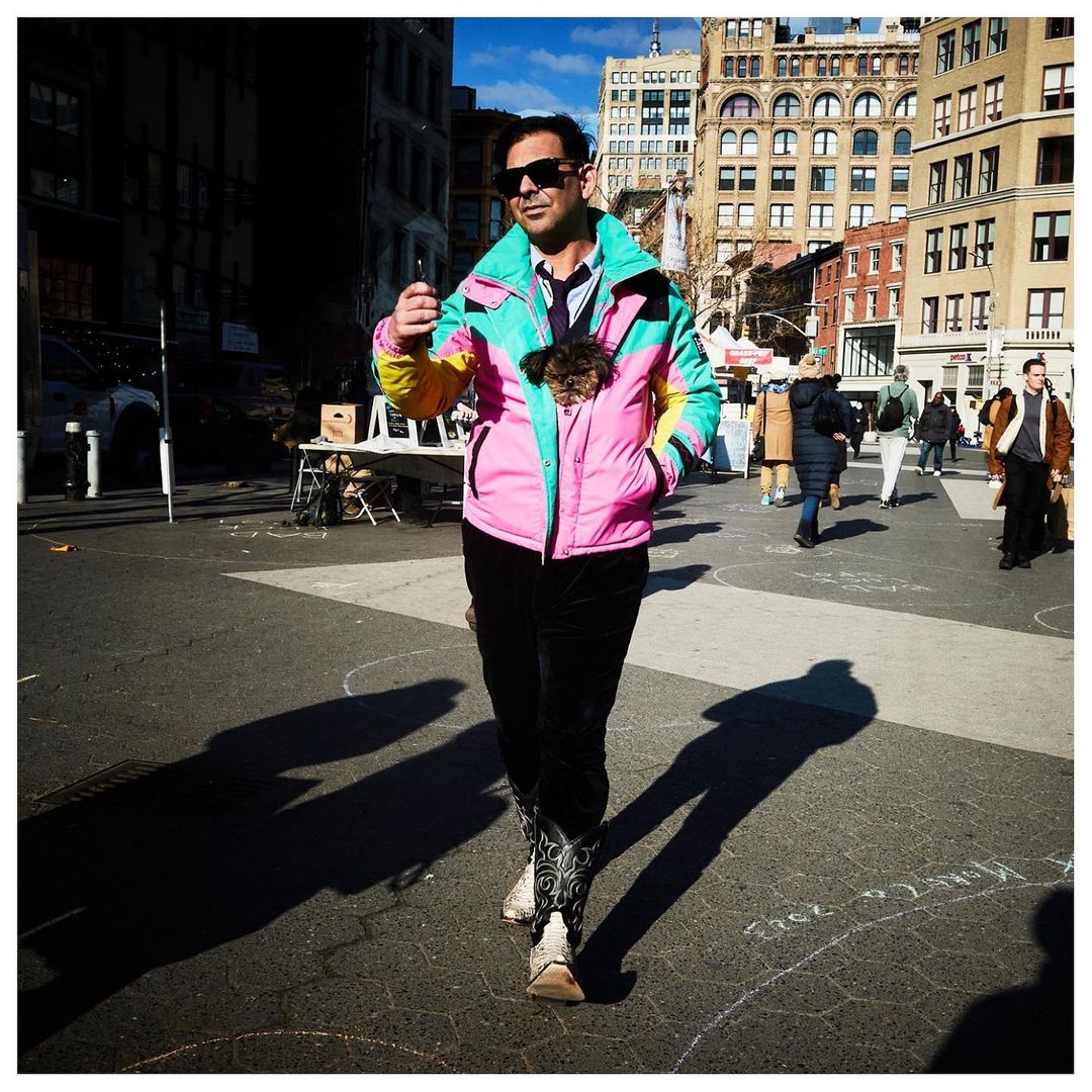 Adult male walks in an urban plaza wearing a colorful jacket with a small dog inside. Buildings and pedestrians are in the background.