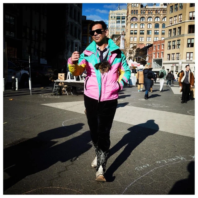 Adult male walks in an urban plaza wearing a colorful jacket with a small dog inside. Buildings and pedestrians are in the background.