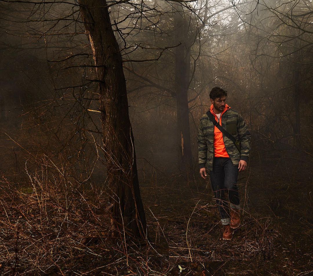 Man walking through a foggy forest wearing a camouflage jacket and an orange shirt with trees and foliage surrounding him.