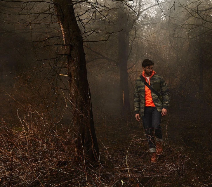 Man walking through a foggy forest wearing a camouflage jacket and an orange shirt with trees and foliage surrounding him.