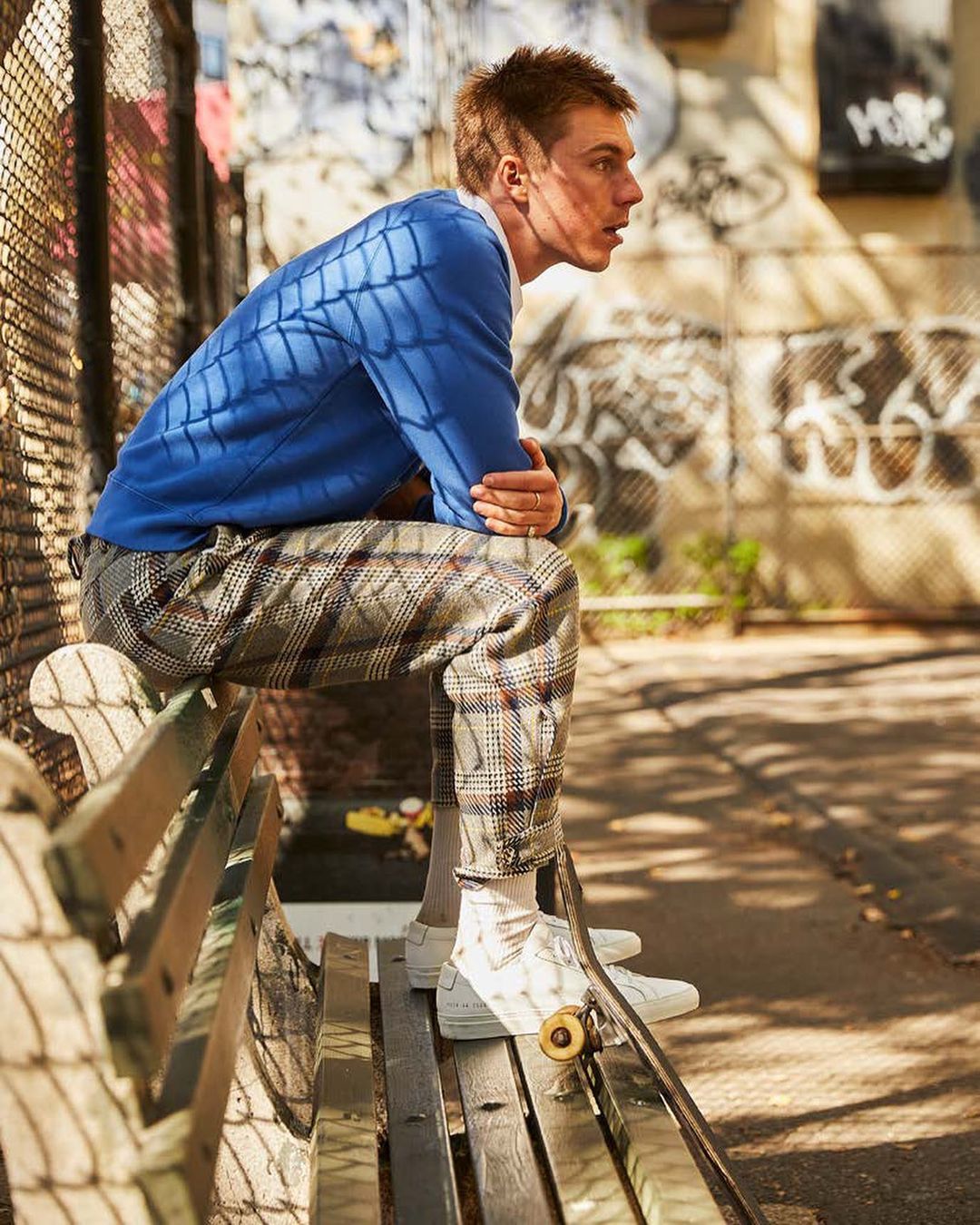 A young adult man sits on a park bench in an urban setting, wearing a blue sweater and plaid pants, next to a skateboard.
