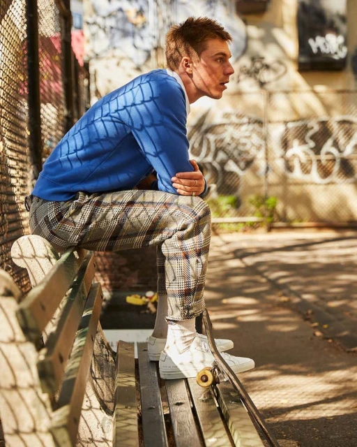 A young adult man sits on a park bench in an urban setting, wearing a blue sweater and plaid pants, next to a skateboard.