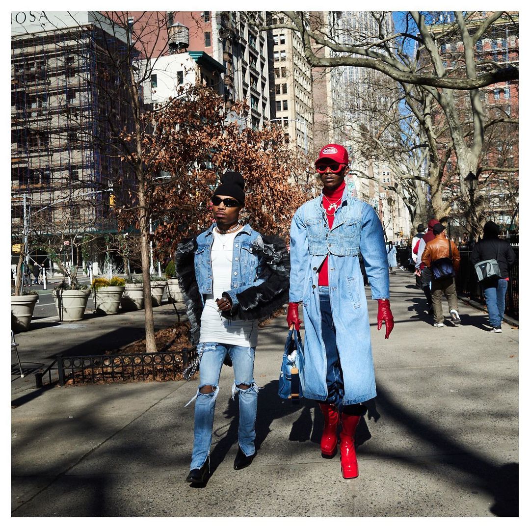 Two men in denim outfits pose on a sunny city street with trees and buildings in the background, showcasing fashion and style.