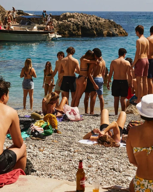 Sun-kissed beach scene with a group of people enjoying the summer, swimming, and socializing near a boat and rocky coastline on a bright day.