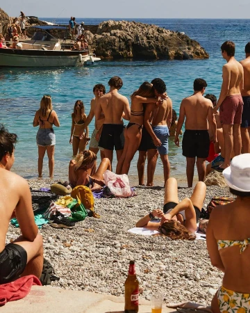 Sun-kissed beach scene with a group of people enjoying the summer, swimming, and socializing near a boat and rocky coastline on a bright day.