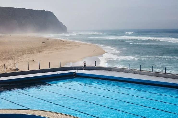 Person standing on the edge of a coastal swimming pool with ocean, beach and mountainous landscape in the background.