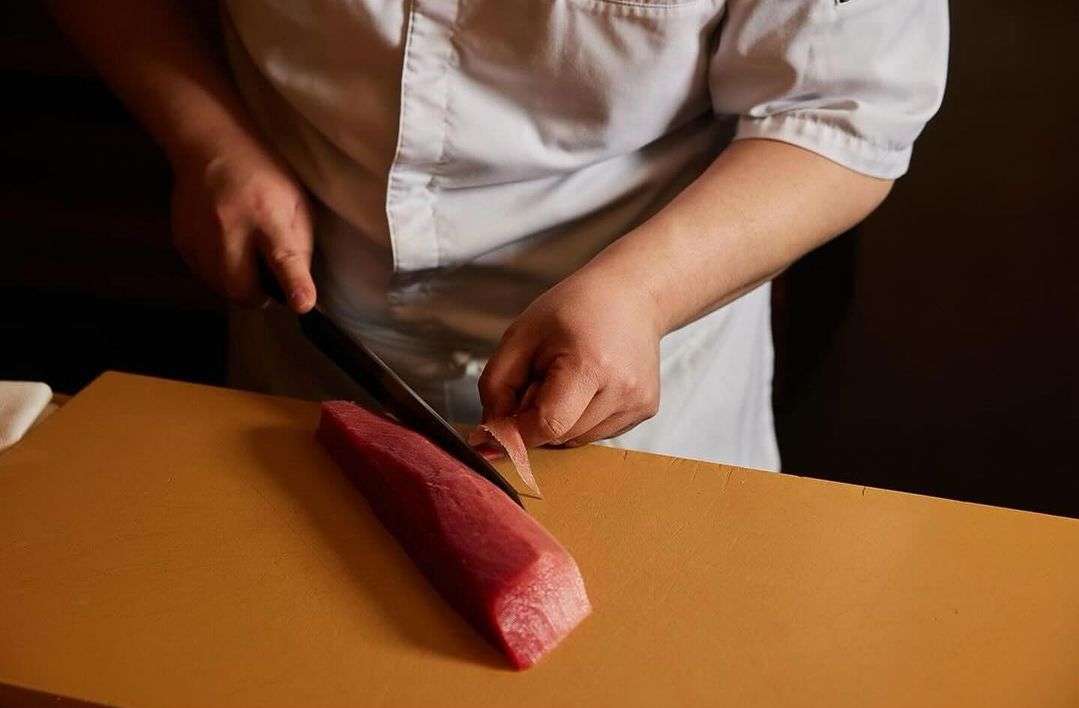 Chef in a white coat meticulously slices a long piece of raw tuna on a wooden cutting board with a sharp knife, indoors.