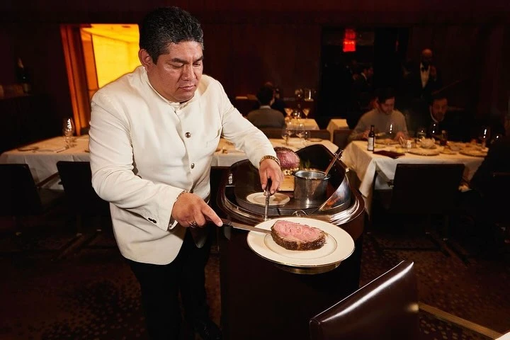 Server in a restaurant cutting a slice of roast beef from a carving station onto a plate, surrounded by diners and elegant decor.