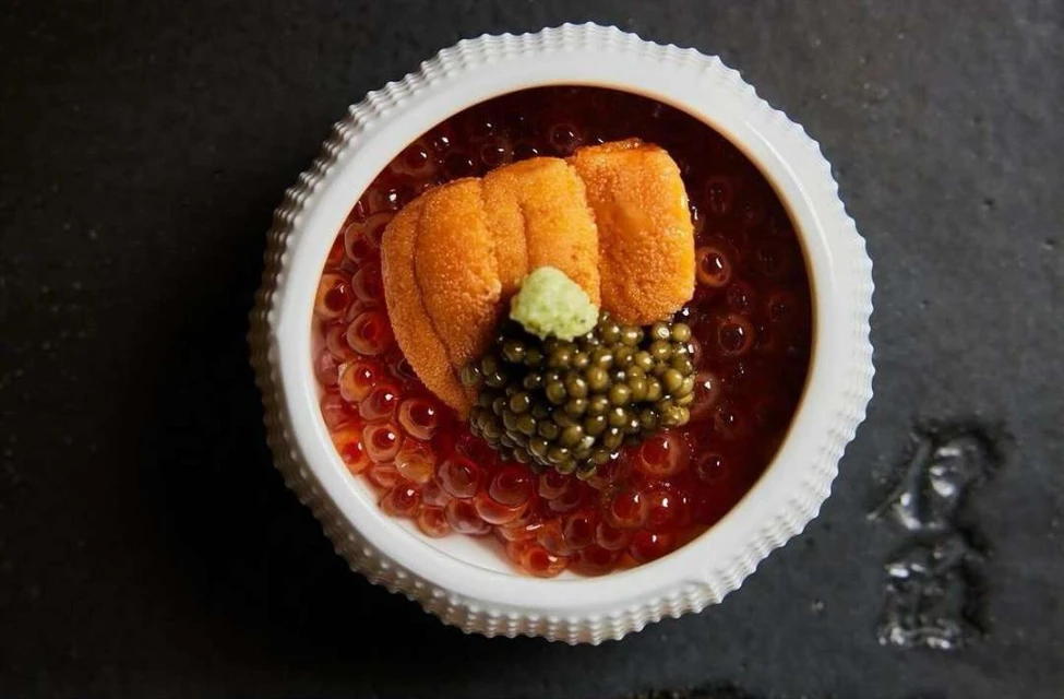 Overhead shot of white bowl filled with salmon roe, sea urchin, and caviar on a dark slate surface. Food photography, studio lighting.