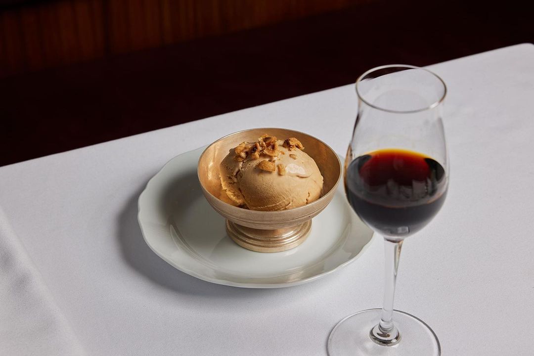 A scoop of tan ice cream sits in a silver bowl, next to a clear glass of red wine, resting on a white tablecloth, indoors.