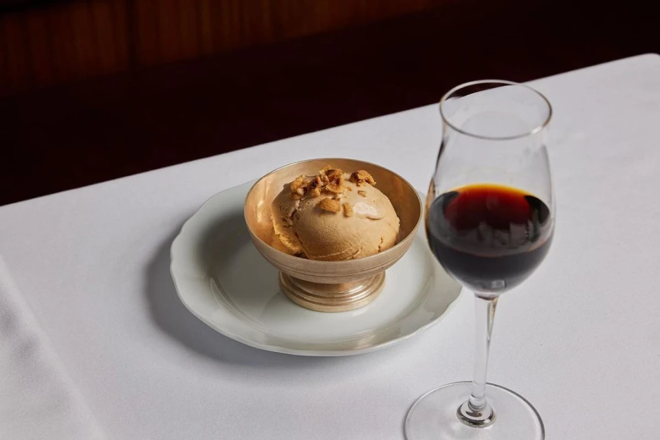 A scoop of tan ice cream sits in a silver bowl, next to a clear glass of red wine, resting on a white tablecloth, indoors.