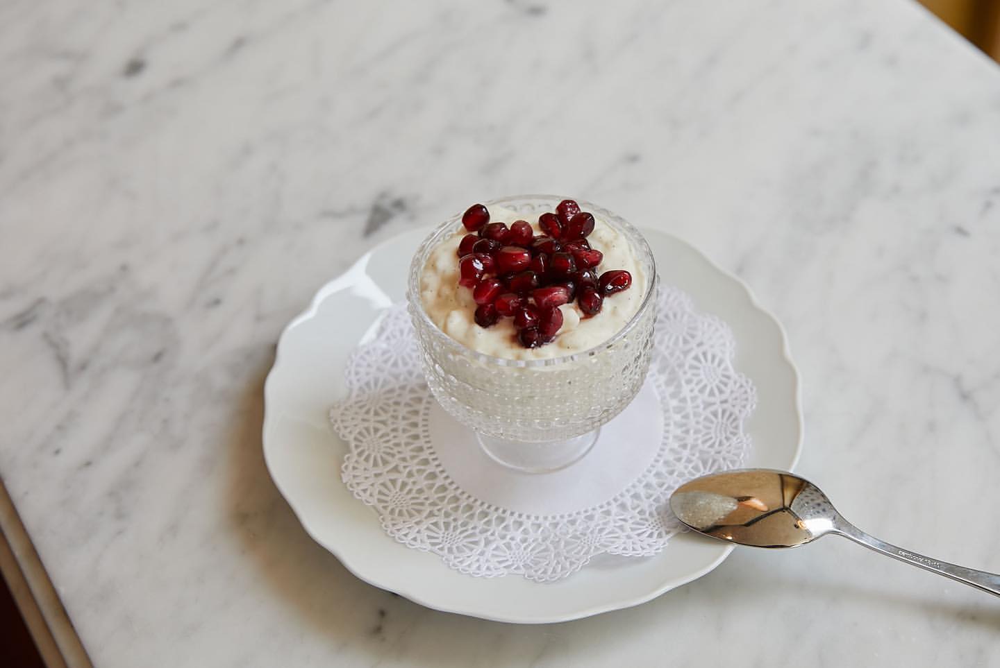 Overhead shot of dessert featuring creamy white pudding in a clear glass, topped with bright red pomegranate seeds, on a white marble table.