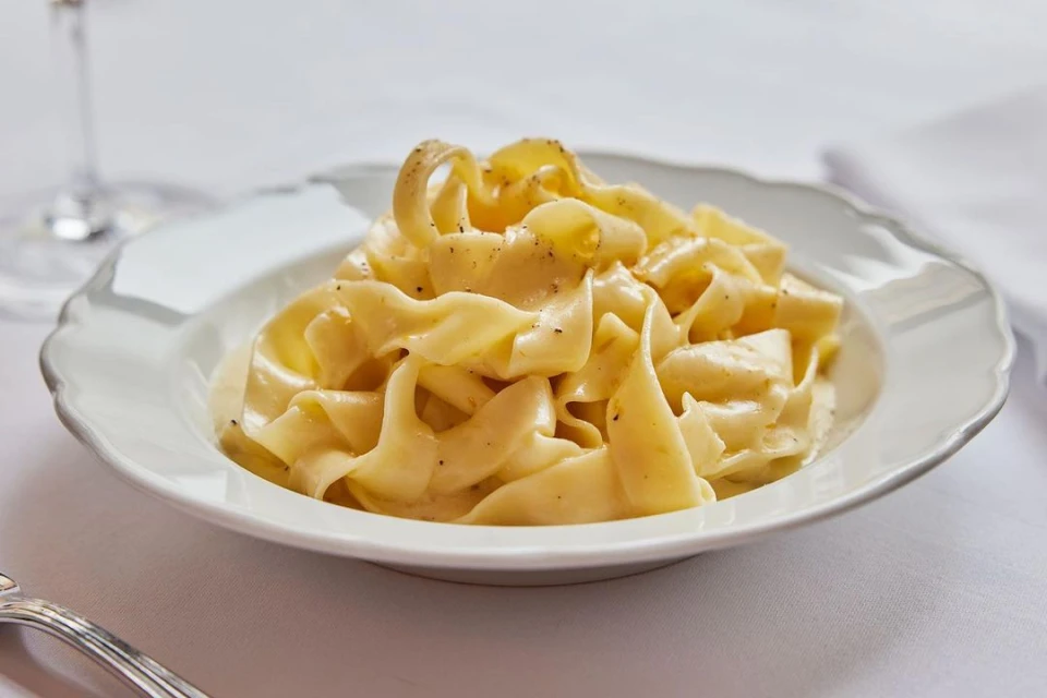 Overhead shot of fresh pappardelle pasta served in a white bowl on a white tablecloth, a partially visible wine glass is in the background.