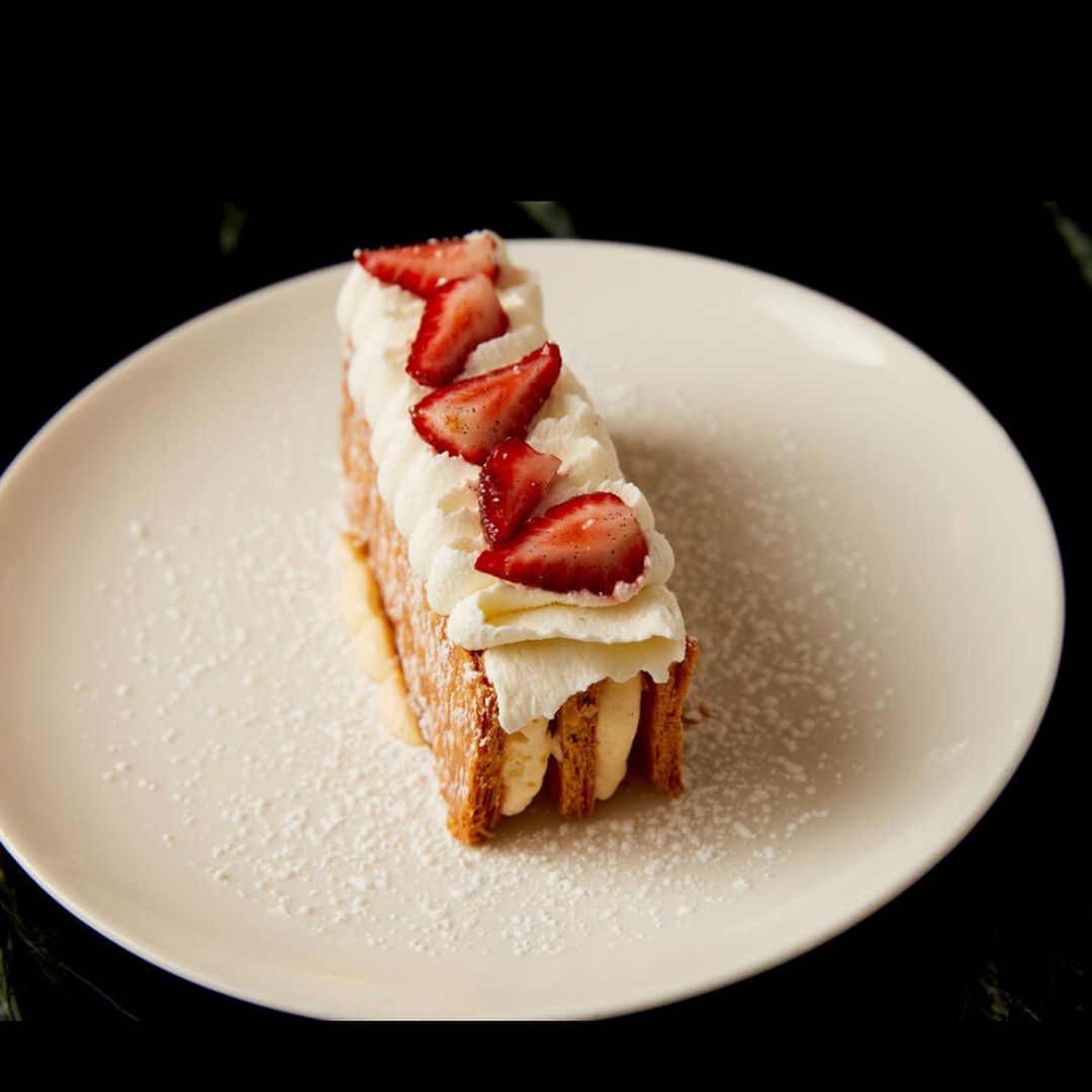 Close-up shot of a mille-feuille pastry topped with whipped cream and fresh strawberries, served on a white plate.