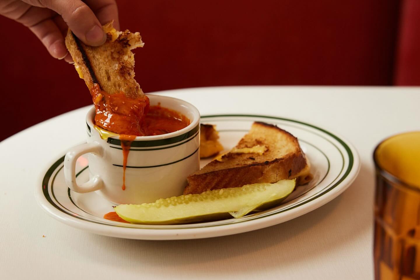 Close-up of hand dipping a grilled cheese sandwich into tomato soup, with a pickle spear and another sandwich slice on a white plate.