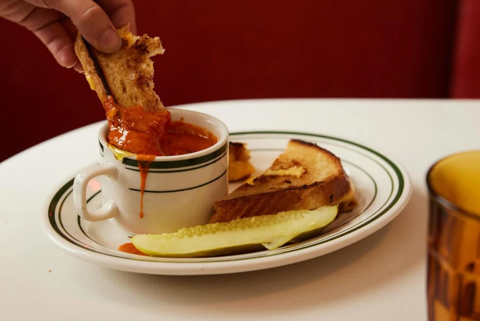 Close-up of hand dipping a grilled cheese sandwich into tomato soup, with a pickle spear and another sandwich slice on a white plate.