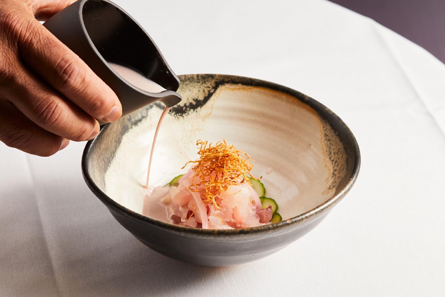 Close-up of a chef pouring sauce from a dark pitcher over a dish of raw fish, garnished with fried shreds, in a black-rimmed ceramic bowl.