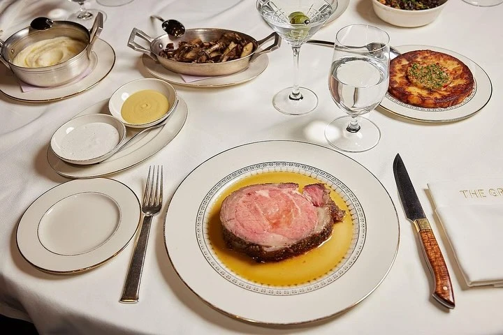 Overhead shot of a restaurant table setting with a steak, mashed potatoes, mushrooms, and cocktails on a white tablecloth.
