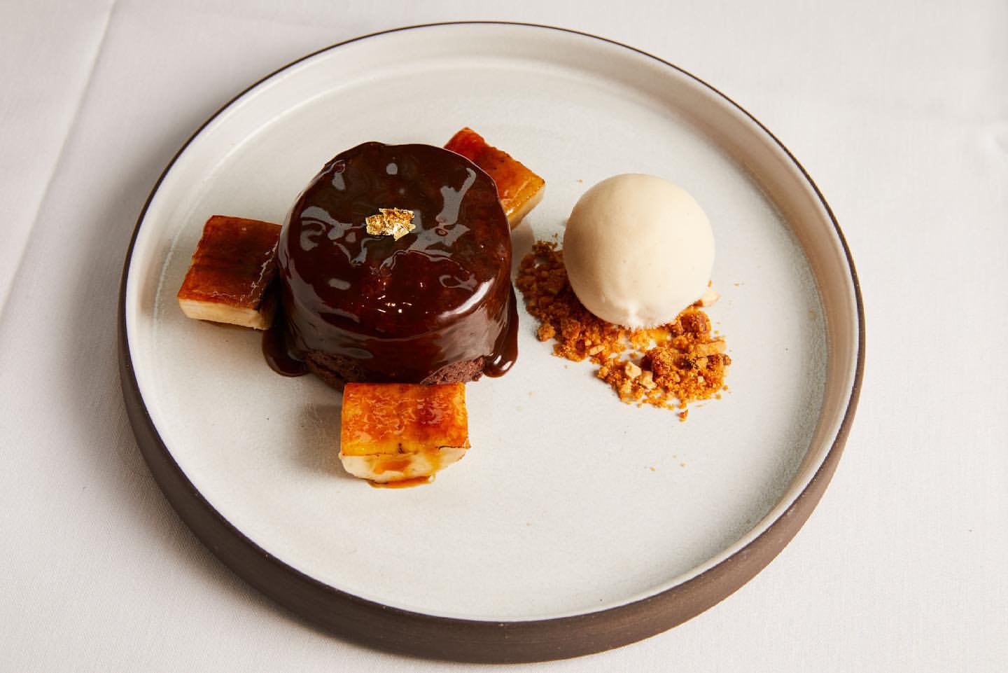 Chocolate lava cake with ice cream and caramel squares on a gray-rimmed white plate, presented on a white tablecloth in a bright setting.