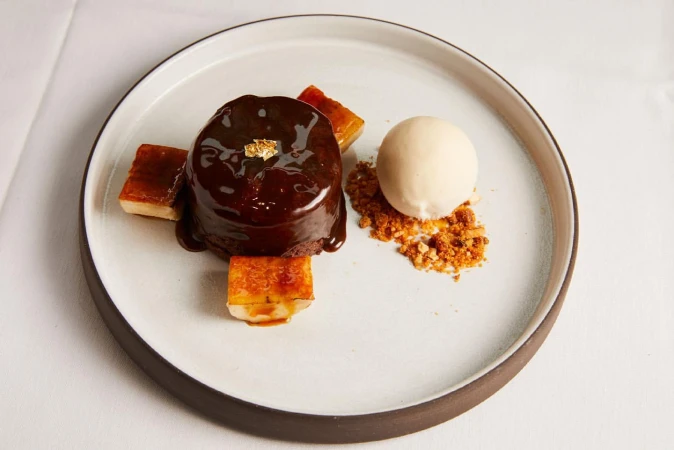 Chocolate lava cake with ice cream and caramel squares on a gray-rimmed white plate, presented on a white tablecloth in a bright setting.