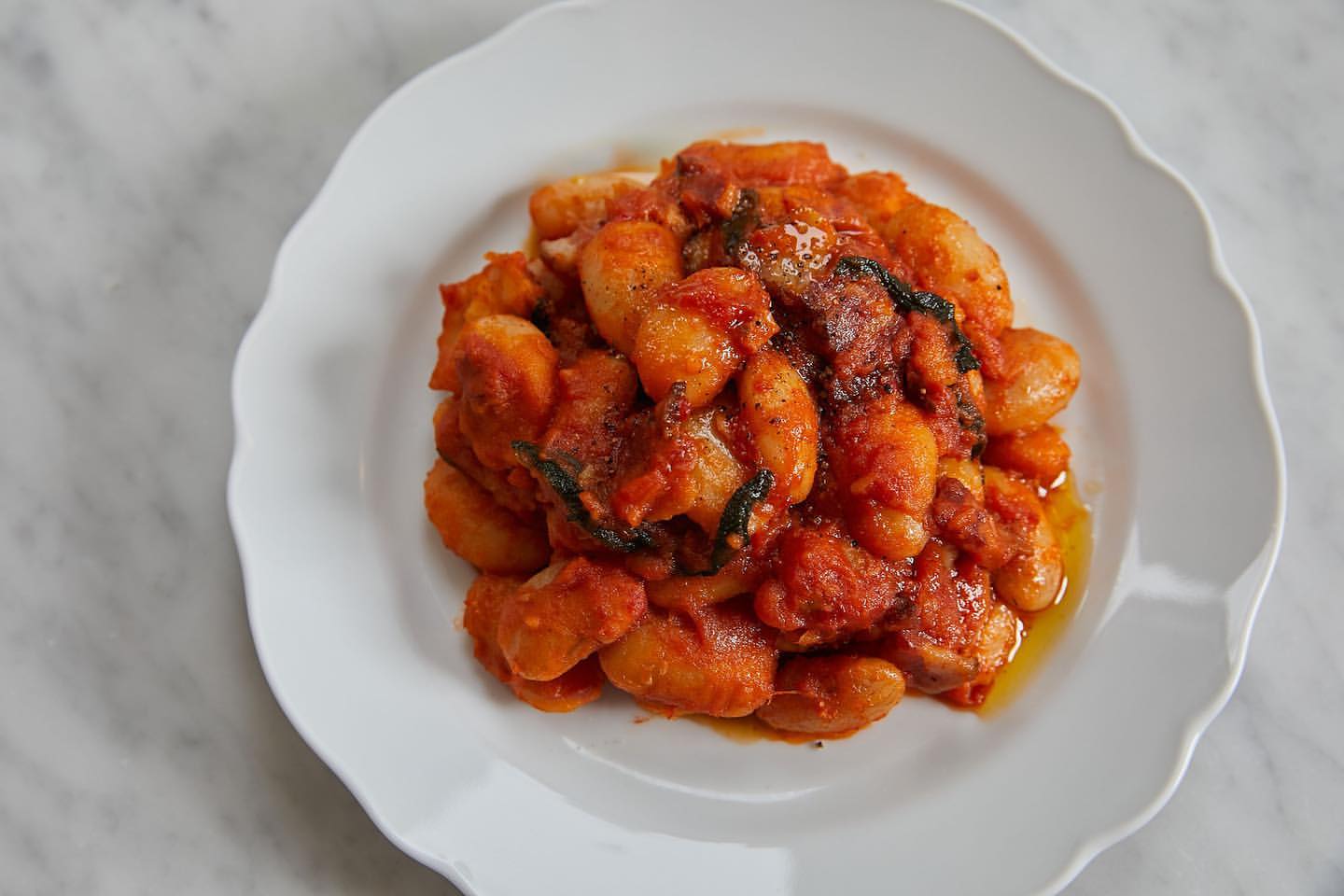 Overhead shot of lima beans in a red tomato sauce, with basil, served on a white porcelain plate over a marble surface.