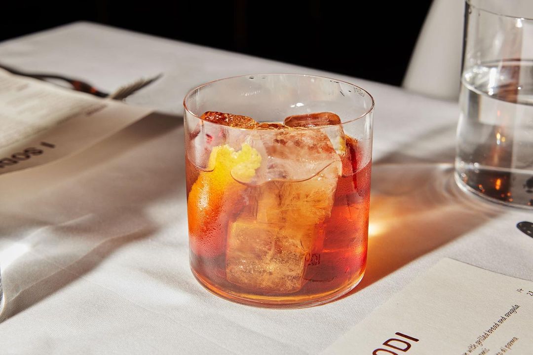 A cocktail drink featuring ice cubes and a piece of orange peel sits on a white tablecloth next to a clear glass of water.