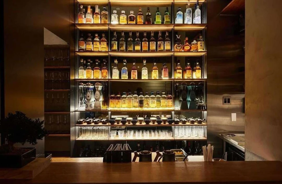 Well-stocked bar featuring shelves of liquor bottles and glassware, illuminated by warm, inviting lighting in a restaurant setting.