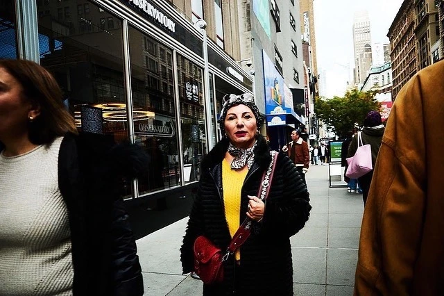Mature woman in yellow sweater and black coat stands on a city sidewalk in New York, with buildings visible in the background.
