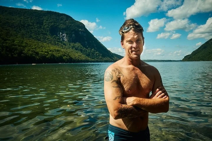 Adult male stands in lake with forested mountains and blue sky with clouds. Arms are crossed and he wears goggles on his head.