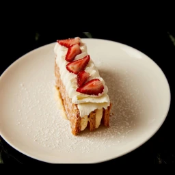 A rectangular pastry topped with whipped cream and strawberry slices on a white plate dusted with powdered sugar.