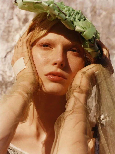 Close-up portrait of a blonde female model with unique headpiece and sheer gloves, posing against a textured stone wall in soft light.