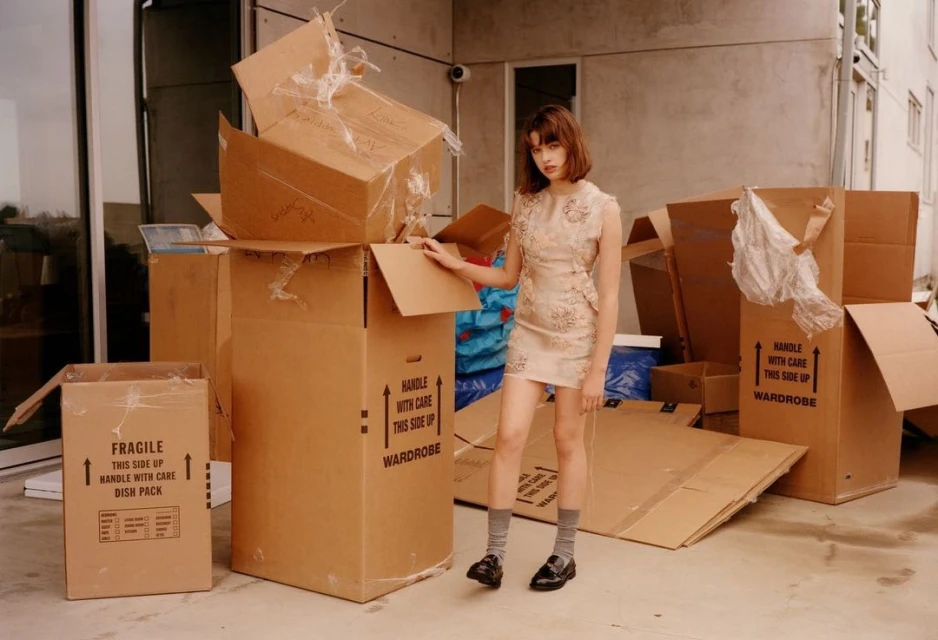 Young adult woman poses in a beige dress amidst stacked cardboard boxes in an outdoor urban setting, editorial fashion photography.