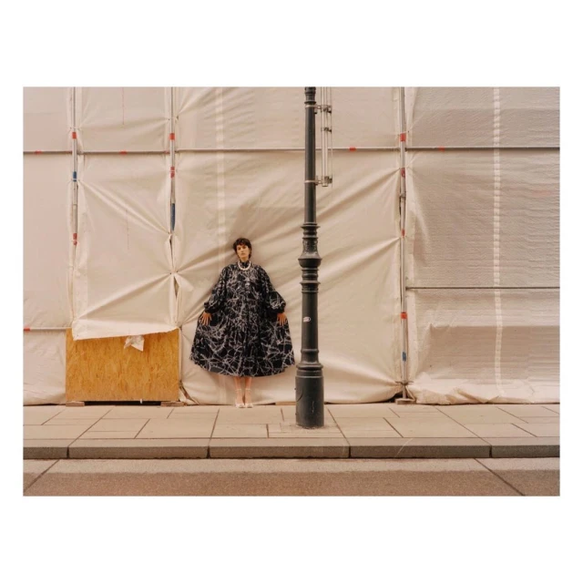 Fashion photography of a woman in a black and white patterned dress standing on a sidewalk against a white construction wall.