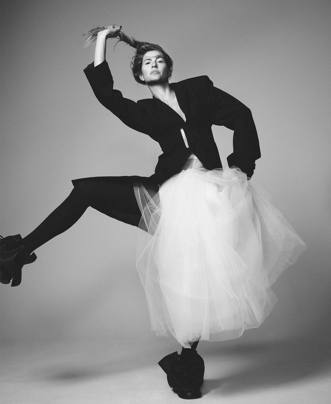A woman in a black blazer and tutu strikes a dynamic pose against a grey background, creating a stark and eye-catching monochrome image.