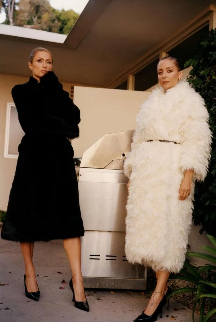 Two women in long coats pose outdoors in front of a home with a grill. One wears black, the other wears white. Both wear black heels.