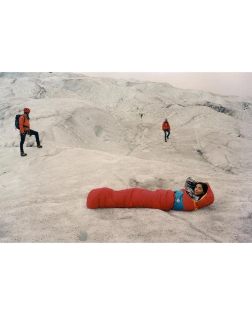 Person in a red sleeping bag reclines on a snowy landscape with three other people visible in the background on a glacier.