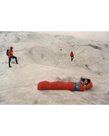 Person in a red sleeping bag reclines on a snowy landscape with three other people visible in the background on a glacier.