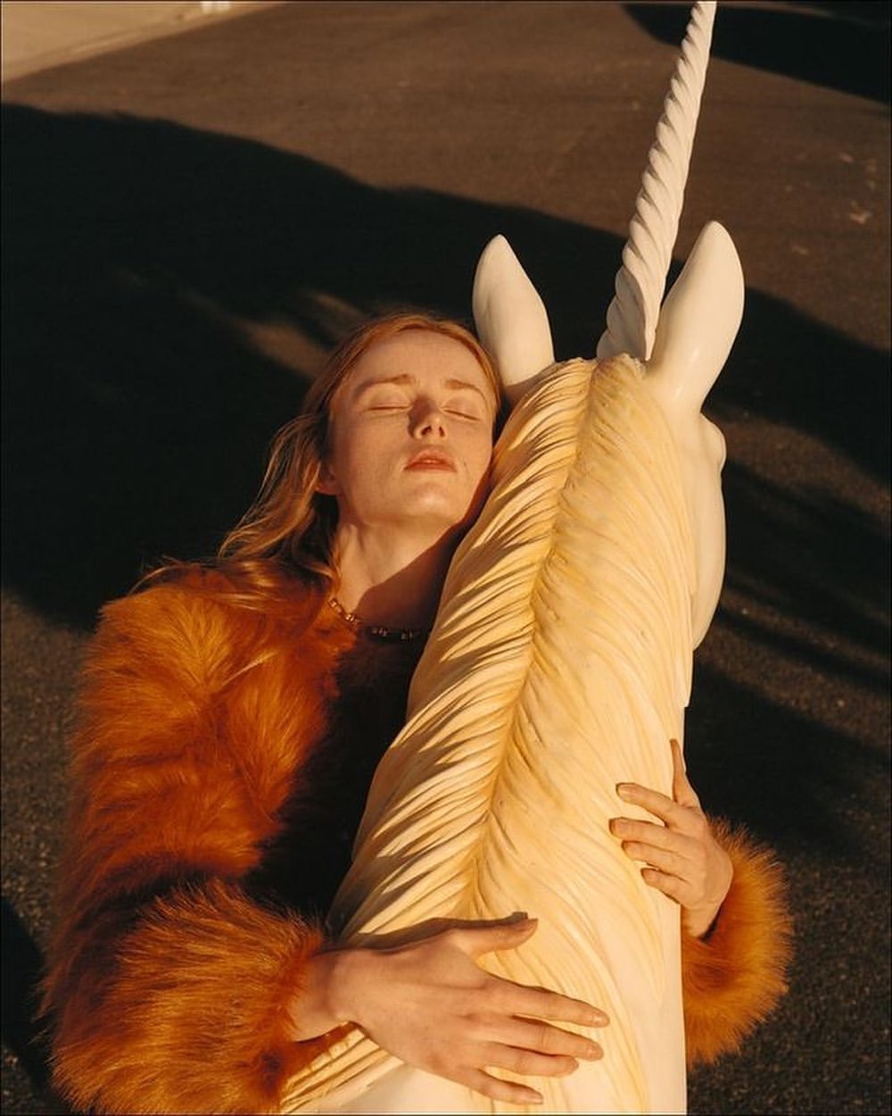 A woman with closed eyes, wearing a fur coat, is embracing a large, sculpted unicorn head on a city street at golden hour.