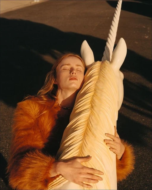 A woman with closed eyes, wearing a fur coat, is embracing a large, sculpted unicorn head on a city street at golden hour.