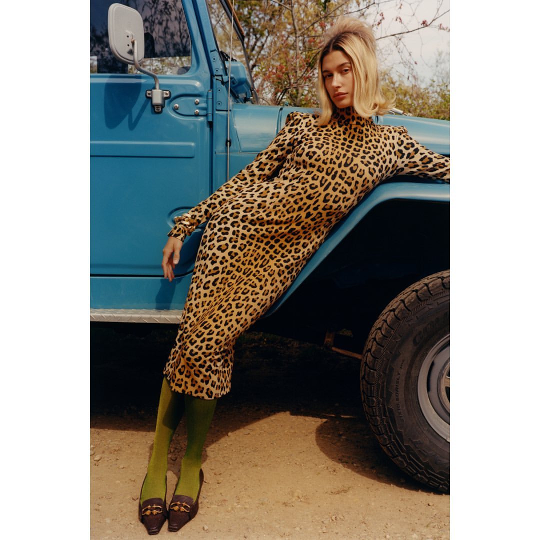 Adult woman poses in a leopard-print dress next to a blue SUV on a dirt road wearing green tights and brown shoes in the daylight.